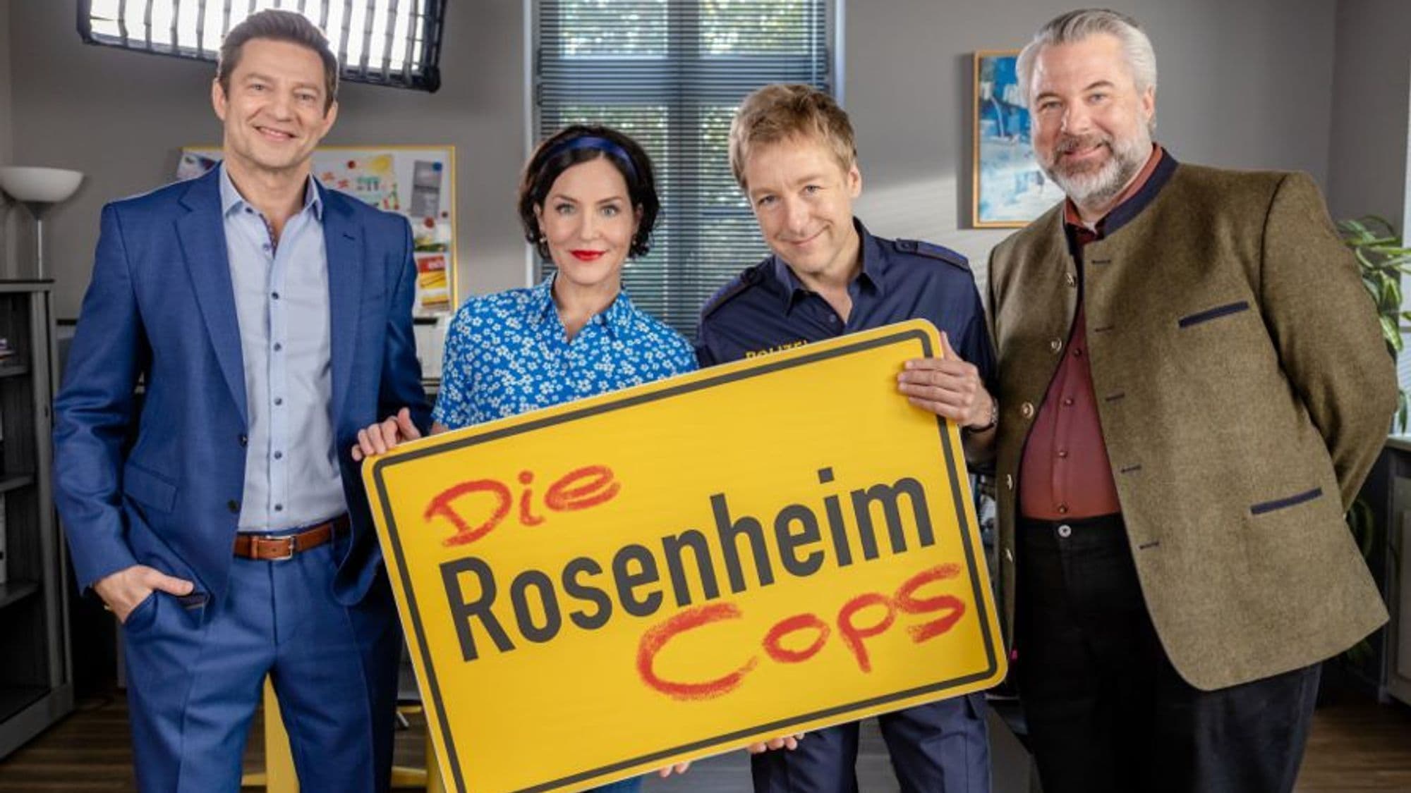 "Die Rosenheim-Cops" ist einer der beliebtesten Krimi-Serien - der altbekannte Cast blickt zufrieden mit Schild in der Hand in die Kamera.